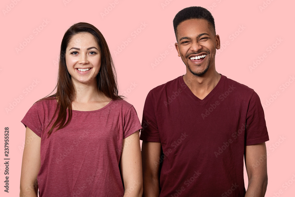Joyful positive African American man and woman smile broadly at camera, have fun together, dressed in casual clothes, isolated over pink wall. Young mixed race couple stand indoor, giggles happily Joyful positive African American man and woman smile broadly at camera, have fun together, dressed in casual clothes, isolated over pink wall. Young mixed race couple stand indoor, giggles happily