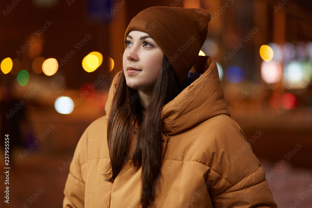 Outdoor shot of thoughtful young woman wears stylish hat and brown jacket, has pensive facial expression, enjoys calm atmosphere in evening city, poses over blurred background. Lifestyle concept Outdoor shot of thoughtful young woman wears stylish hat and brown jacket, has pensive facial expression, enjoys calm atmosphere in evening city, poses over blurred background. Lifestyle concept