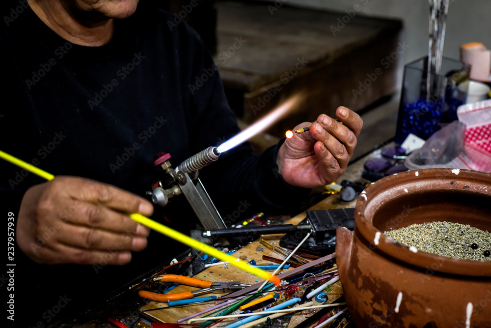 Obraz premium a glassblower man made Handicraft from melted glass in glassblowing workshop. Glass art is shaping at the end of glassblowing pipe. 