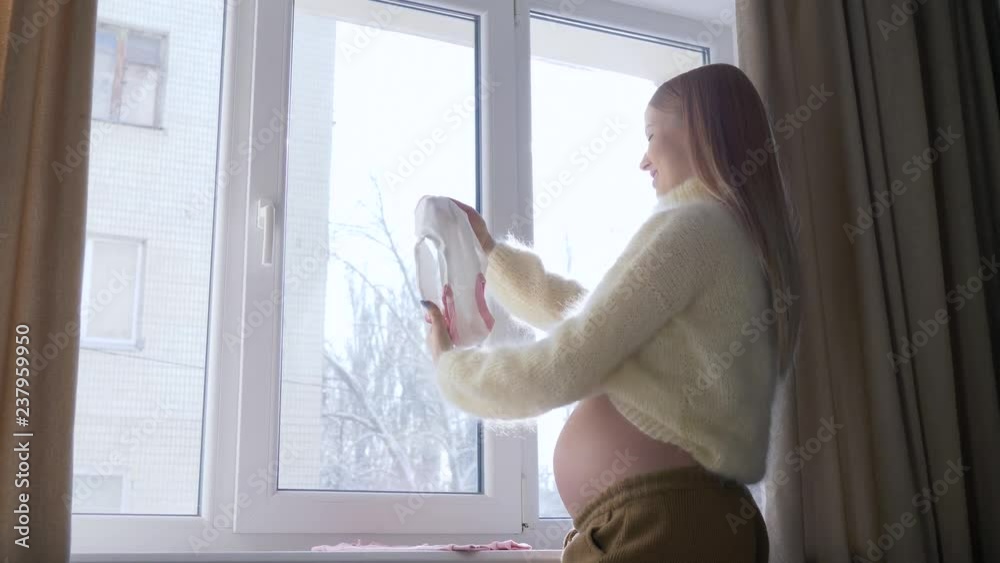 Waiting of baby, young pregnant female with big tummy looks at little children clothes for future baby against window in sunlight on winter Waiting of baby, young pregnant female with big tummy looks at little children clothes for future baby against window in sunlight on winter