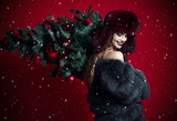 Happy fashion woman in winter hat and fur coat hold decorated xmas tree on her shoulders smiling