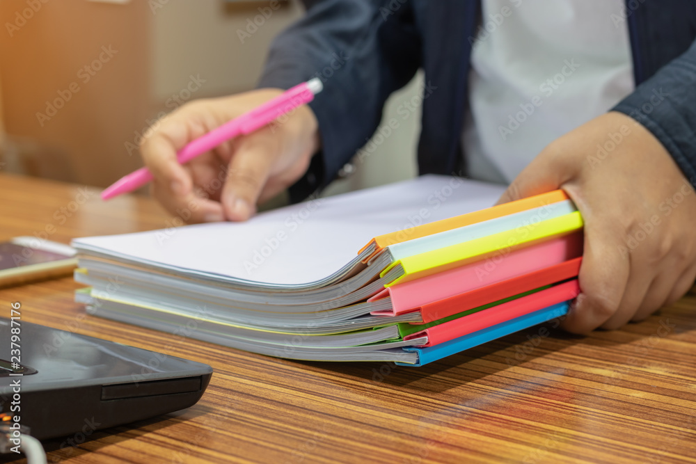 custom made wallpaper toronto digitalTeacher hand is holding pen and unfinished student homework assignments on desk in school office. Paperwork stacked in archive with color plastic slide binder bars. Education and business concept.