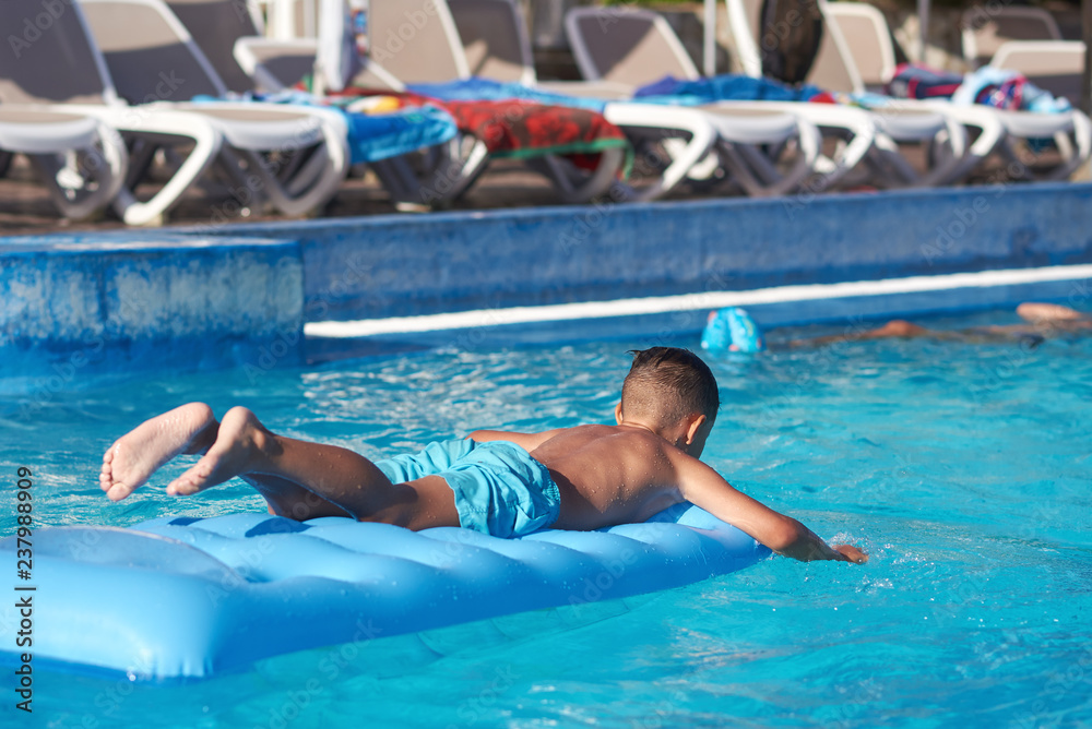 Cute European boy is using blue air mattress, while having fun in hotel’s swimming pool.