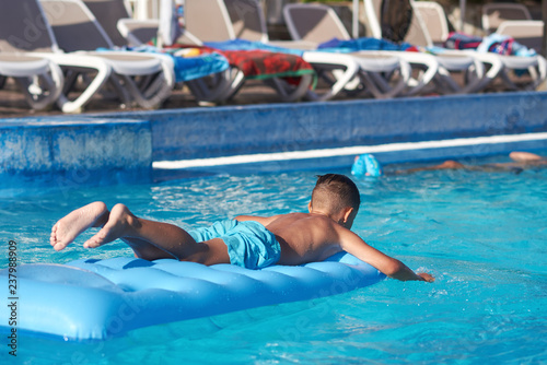 Fototapeta Naklejka Na Ścianę i Meble -  Cute European boy is using blue air mattress, while having fun in hotel’s swimming pool.