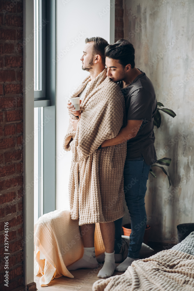 Nice same-sex married couple is standing together at window. Careful and gentle male brunette embracing from the back his woman-like male friend wrapped in soft blanket