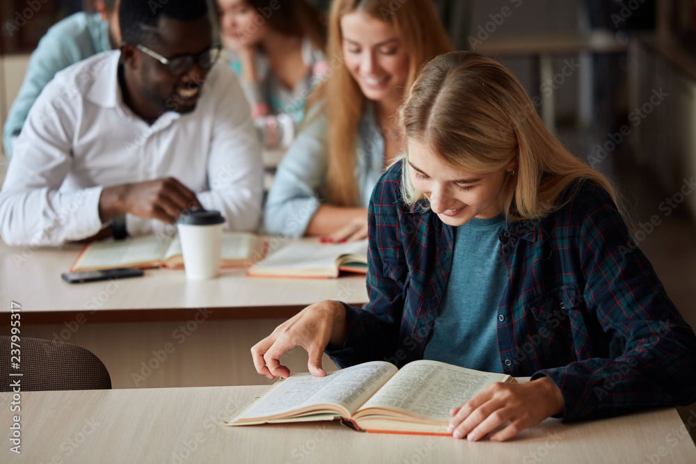 Smiling attractive blonde girl makes some notes while reading book in college library, looking at camera, sitting at desk with the other students on background Smiling attractive blonde girl makes some notes while reading book in college library, looking at camera, sitting at desk with the other students on background