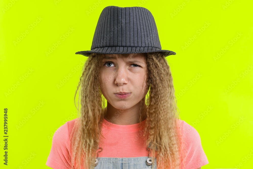 Portrait of a pretty funny teen girl with a pensive face, blonde girl with curly hair in hat with thoughtful expression, presses lips together, yellow studio background Portrait of a pretty funny teen girl with a pensive face, blonde girl with curly hair in hat with thoughtful expression, presses lips together, yellow studio background