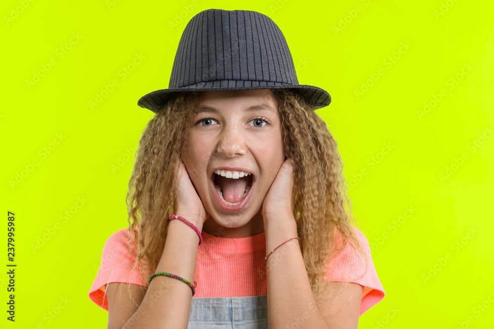 Surprised happy teen girl with curly hair in hat looking at the camera with open mouth over yellow studio background