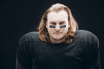 Portrait of determined caucasian blonde American football sportsman in black uniform with painted black stripes under his eyes on his face posing against black background