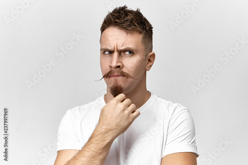 Horizontal shot of handsome young male dressed in casual wear frowning suspiciously and stroking his goatie beard, thinking about something or scheming tricky plan. Human feelings and emotions photo