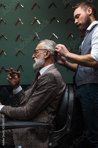 Barber making male haircut. Hairdresser cutting hair of his rich client, dressed in expensive stylish man s suit of clothes and holding a glass of alcoholic beverage