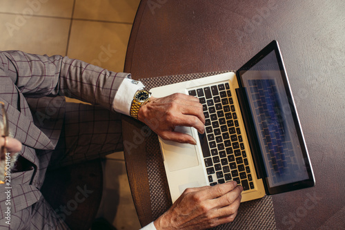 High angle view of male hands with gold wristwatch, dressed in grey expensive suit, working on laptop over red wood table background