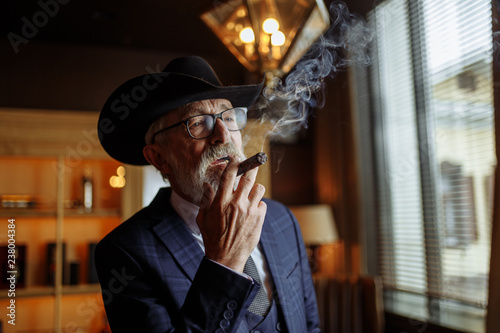 Stylish old-aged dandy in wide brimmed hat and rich dark blue mens suit smoking cigar indoor, standing near the bar counter with alcohol drinks.