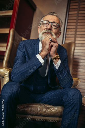 Serious concentrated old male creative artist wears glasses and formal blue suit, with wrinkled and experienced face, remembering remarkable life s moments while sitting in the restautant