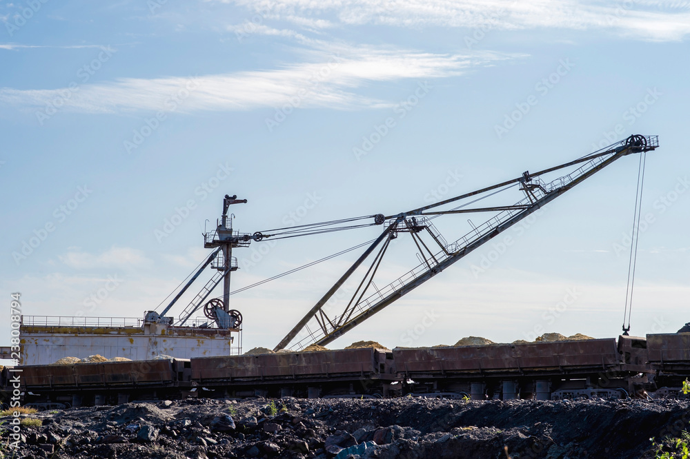 A huge excavator overloads in the dumps rock from the unloaded train from the mine against a clear blue sky. Concept: mining and environmental problems