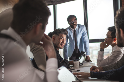 deadline, technology and people concept - creative team of multiracial men, sitting round the desk in diverse casual and formal clothes talking over new project in office meeting room photo