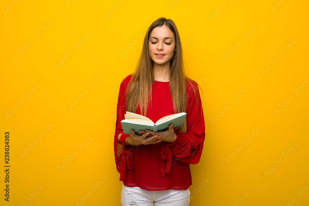 Young girl with red dress over yellow wall holding a book and enjoying reading Young girl with red dress over yellow wall holding a book and enjoying reading