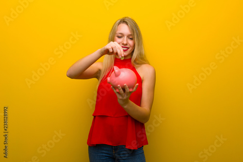 Young girl with red dress over yellow wall taking a piggy bank and happy because it is full