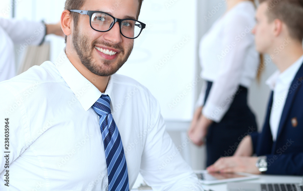 Business people at meeting in office. Focus at cheerful smiling bearded man wearing glasses. Conference, corporate training or brainstorming of people group. Success and negotiation concept Business people at meeting in office. Focus at cheerful smiling bearded man wearing glasses. Conference, corporate training or brainstorming of people group. Success and negotiation concept