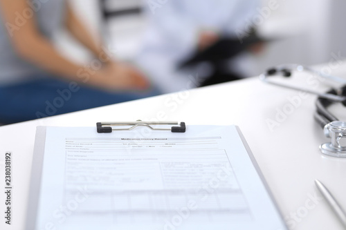 Stethoscope, clipboard with medical form lying on hospital reception desk with laptop computer and busy doctor and patient communicating at the background. Medical tools at doctor working table