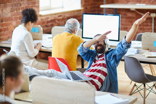 Fotografie Young ecstatic office manager in headphones making dab gesture while sitting by