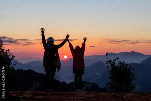 Silhouettes of happy mother and daughter looking morning sunrise on peak of mountain.