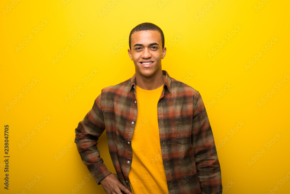 Fototapeta premium Young african american man on vibrant yellow background posing with arms at hip and smiling
