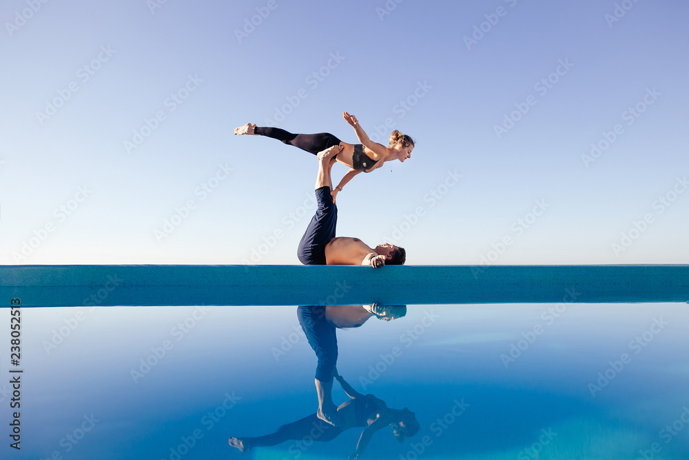 Acro yoga concept. Pair yoga. Couple of young sporty people practicing yoga lesson with partner, man and woman in yogi exercise, arm balance pose, working out by pool, above beach, against blue sky Acro yoga concept. Pair yoga. Couple of young sporty people practicing yoga lesson with partner, man and woman in yogi exercise, arm balance pose, working out by pool, above beach, against blue sky