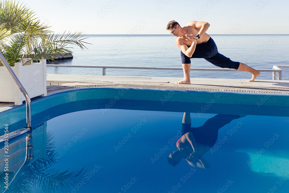 Young attractive man practicing yoga doing Parivrtta Parsvakonasana exercise, Revolved Side Angle pose, working out by the pool, above the beach, relaxing against blue sky. Health and beauty concept