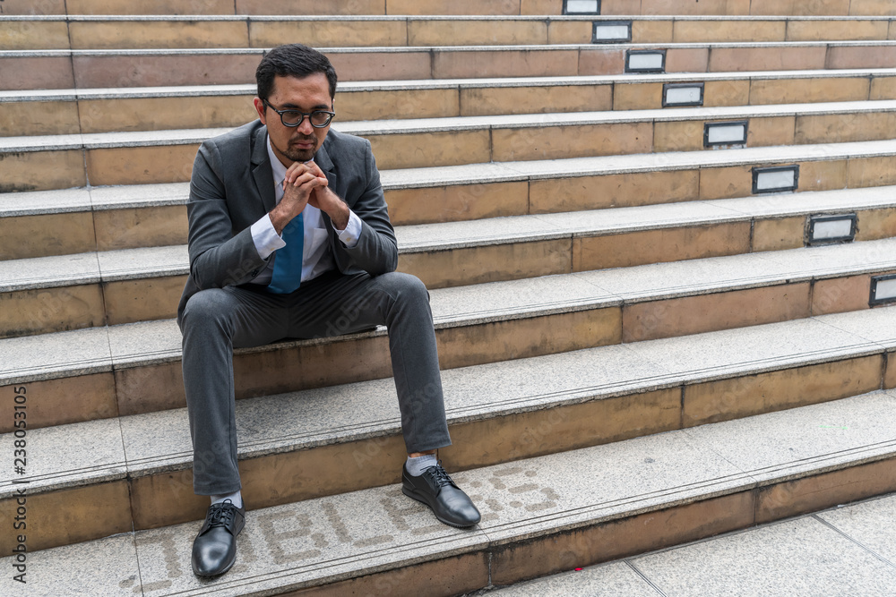 A sad looking young businessman sitting at the staircase looking down with stressed and depressed look gesture with the word A sad looking young businessman sitting at the staircase looking down with stressed and depressed look gesture with the word