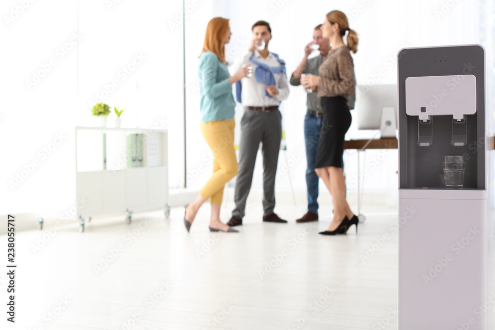 Modern water cooler with glass and blurred office employees on background. Space for text Modern water cooler with glass and blurred office employees on background. Space for text