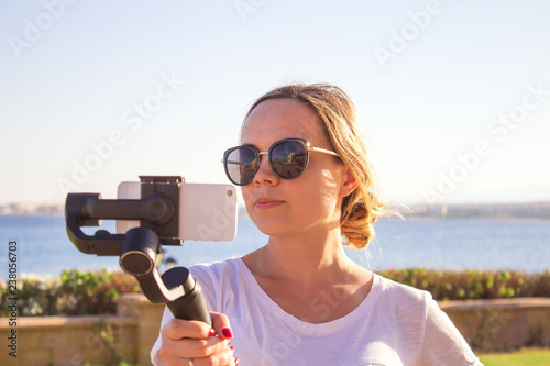 Video blogger girl. Operator with action camera on stabilized grip with gimbal. Happy young woman shoots 4k footage on event outdoor