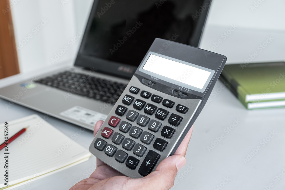 White screen calculator in men's hands There is a laptop and pencil on notebook on the white desk as a backdrop. White screen calculator in men's hands There is a laptop and pencil on notebook on the white desk as a backdrop.