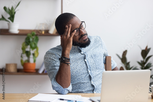Serious african millennial businessman thinking cogitating about business issues. Frustrated black entrepreneur searching problem solution sitting alone on chair at modern office desk looking away