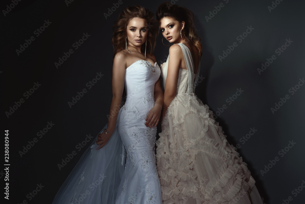 Two beautiful brides with perfect make up and hairstyle wearing luxurious wedding dresses and splendid earrings. Isolated on dark grey background. Studio shot.