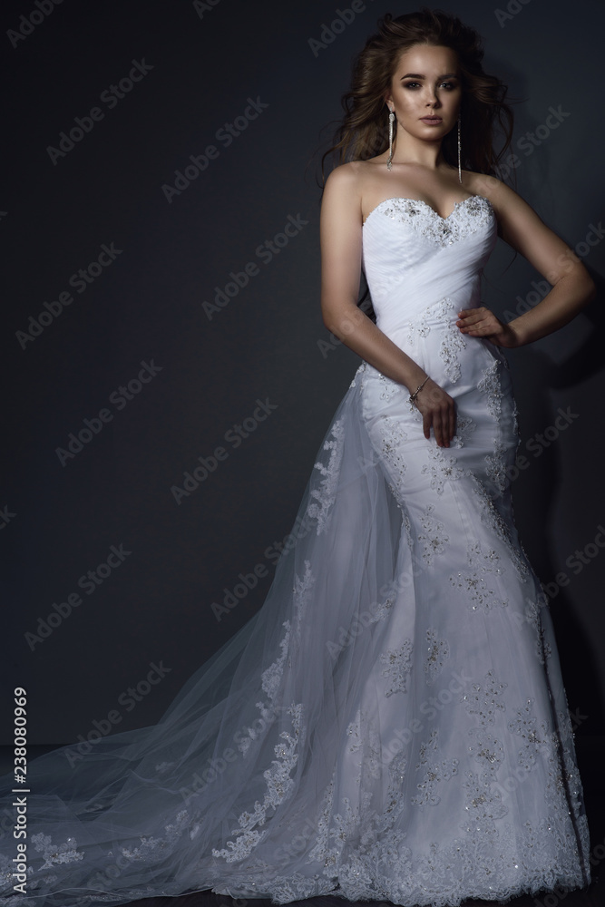 Beautiful young model with perfect make up and blowing hair wearing luxurious mermaid white lace wedding dress with long veiling train. Isolated on grey background. Text space.