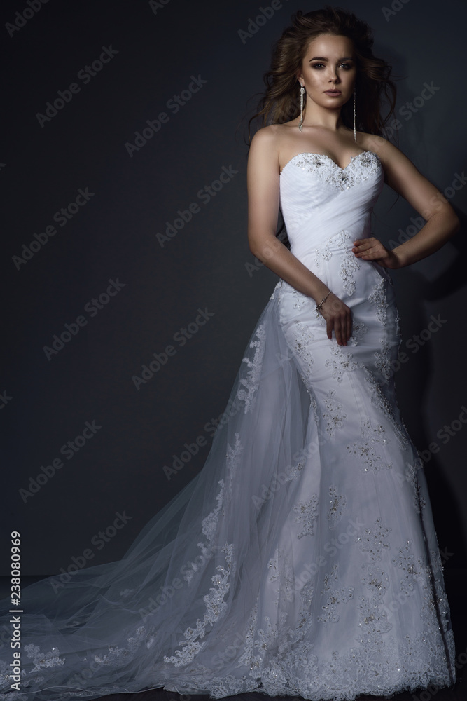 Beautiful young model with perfect make up and blowing hair wearing luxurious mermaid white lace wedding dress with long veiling train. Isolated on grey background. Text space. Beautiful young model with perfect make up and blowing hair wearing luxurious mermaid white lace wedding dress with long veiling train. Isolated on grey background. Text space.