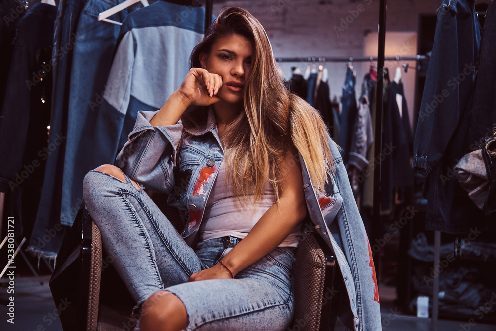 A seductive girl wearing a distressed jeans and denim jacket looking at a  camera while sitting on a chair in the fitting room of a clothing store.  Stock Photo | Adobe Stock
