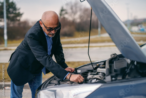 Bearded senior adult man standing in front of opened hood of his car and trying to fix engine.