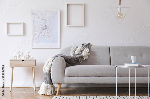 Minimalistic scandinavian interior with design sofa, coffee table, cozy blanket, poster map and mock up photo frames. White background walls and modern triangle lamp. 