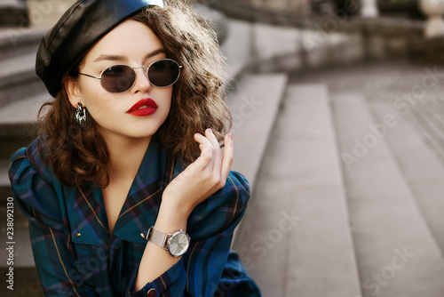 Outdoor fashion portrait of young beautiful fashionable girl wearing trendy sunglasses, wrist watch, earrings, leather beret, blue checkered dress, posing in street, on stairs. Copy, empty space