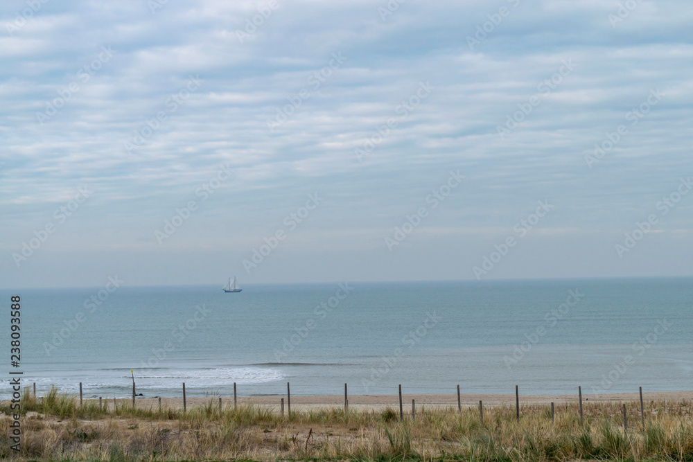 Seascape with coastline and calm blue sea with a sailboat on the horizon line. A deserted beach on the North Sea coast. Sand dunes on the coast with seaside grass Ammophila arenaria. 