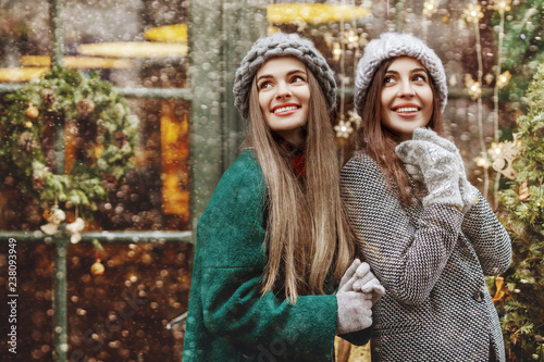 Outdoor portrait of two young beautiful fashionable happy smiling surprised girls posing at festive Cristmas fair. Models looking up, wearing stylish winter clothes. Copy, empty space for text