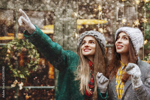 Happy smiling surprised girl pointing up to her friend. Models looking up, wearing stylish winter clothes. Snowfall, festive Christmas background. 