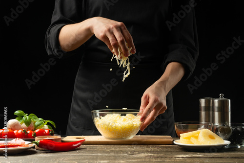 The chef prepares mozzarella cheese, cheddar for Italian pizza, pasta. On a black background, freezing in motion. A concept of tasty and healthy food. Design text, recipe book