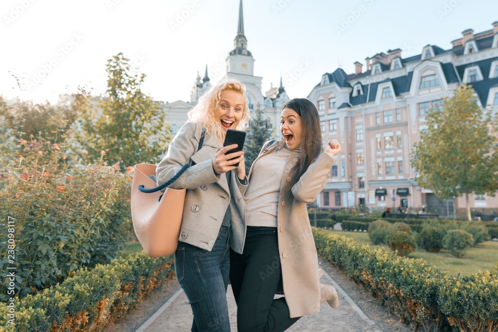 Obraz premium Two young women having fun, looking at the smartphone laughing, sunny autumn day, city background, golden hour