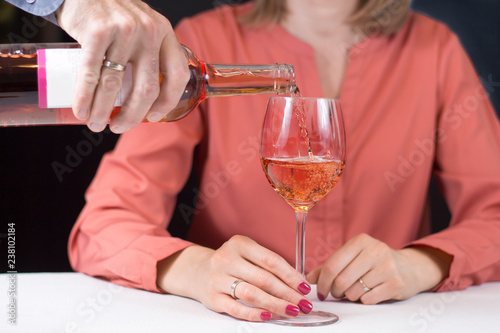 Kobieta siedzi przy stole i trzyma w dłoni kieliszek do wina. Męska dłoń nalewa wina z butelki do kieliszka.