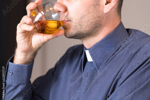 Ksiądz w koloratce i szarej koszuli pije whisky z lodem ze szklanki.