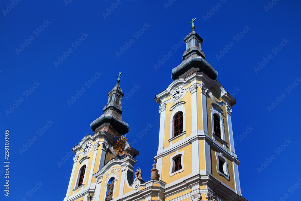 Low-angle view of two yellow and white clock towers of sunlit Baroque Minorite church against clear blue sky, downtown Heroes' Square in Hungarian city Miskolc, Borsod county Central / Eastern Europe