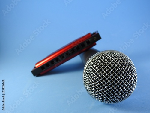 A dynamic handheld vocal microphone and a red 10-hole diatonic harmonica. The head of the microphone, in the foreground, is focused and the harmonica is out of focus. Blue background.